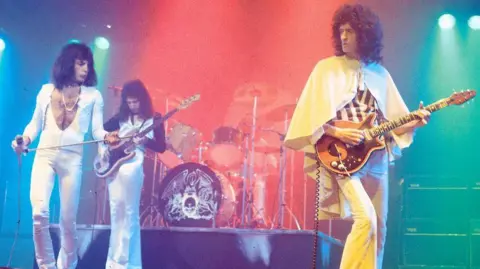 Getty Images Freddie Mercury , John Deacon and Brian May performing live on stage. Freddie is wearing a white jump suit, open over his chest and is wearing a gold chain. He is holding a microphone. Behind him John Deacon has a white and black jumpt suit, holding a guitar, and on the right of the picture is Brian May, in a flamboyant yellow outfit, playing his guitar. 