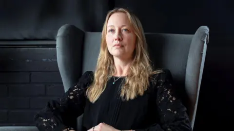 Ali Smith Katie Ward sits in a large grey armchair looking out of frame. She is blonde and wears a black top.