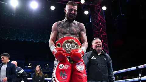 Getty Images Lewis Crocker has a red belt around his waist and his red boxing gloves and shorts on. He has tattoos across the upper half of his body and is screaming in the picture after winning the fight.  His father, who has short grey hair and a black coat, is standing next to him. There are people standing in the ring behind him.
