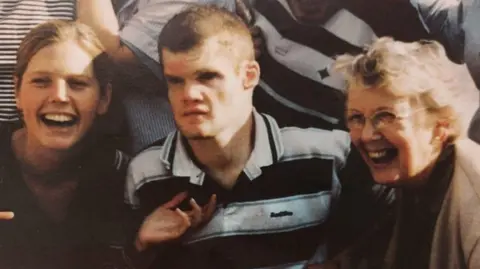 Family Handout A man wearing a black and white striped top. He is sat in between two women.