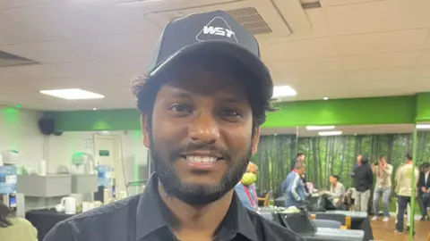 James Patient/BBC Kumar Kuppusamy has dark hair, a beard and is wearing a black shirt. He has a black cap on his head. Behind him, people are gathered in a media room with a green wall.
