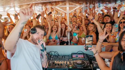 Tom Hall A man who is a DJ holding a microphone to his mouth. He has a crowd of young people gathered behind him who are partying to his music. They are all on a yacht. The young men and women are wearing swimwear. 