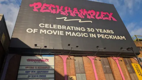 George Walker/BBC A sunlit image of the front of the Peckhamplex cinema including a pink graffiti style logo for Peckhamplex on a black background which states the cinema is celebrating 30 years of movie magic in Peckham