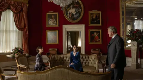 Rory Mulvey 2025 FOCUS Downton Abbey characters Cora, Lady Mary and Lord Granthan are in a red ornately designed room talking to one another. There are portrait paintings in golden frames on the wall and two sofas in green velvet in the centre of the room. Lord Granthan wears a grey suit, while Cora and Lady Mary wear tea dresses.