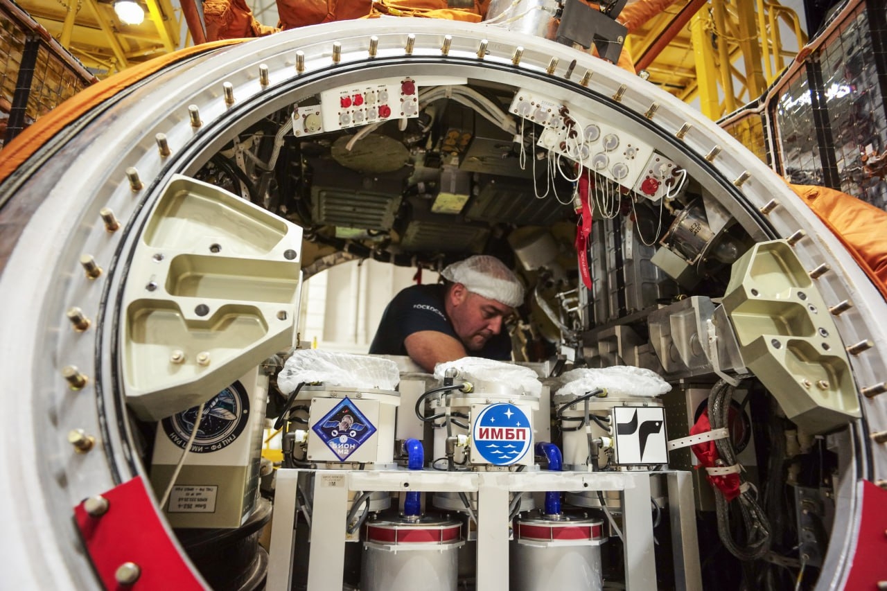A man crouches in the dome of the Bion-M no. 2 module next to storage containers.
