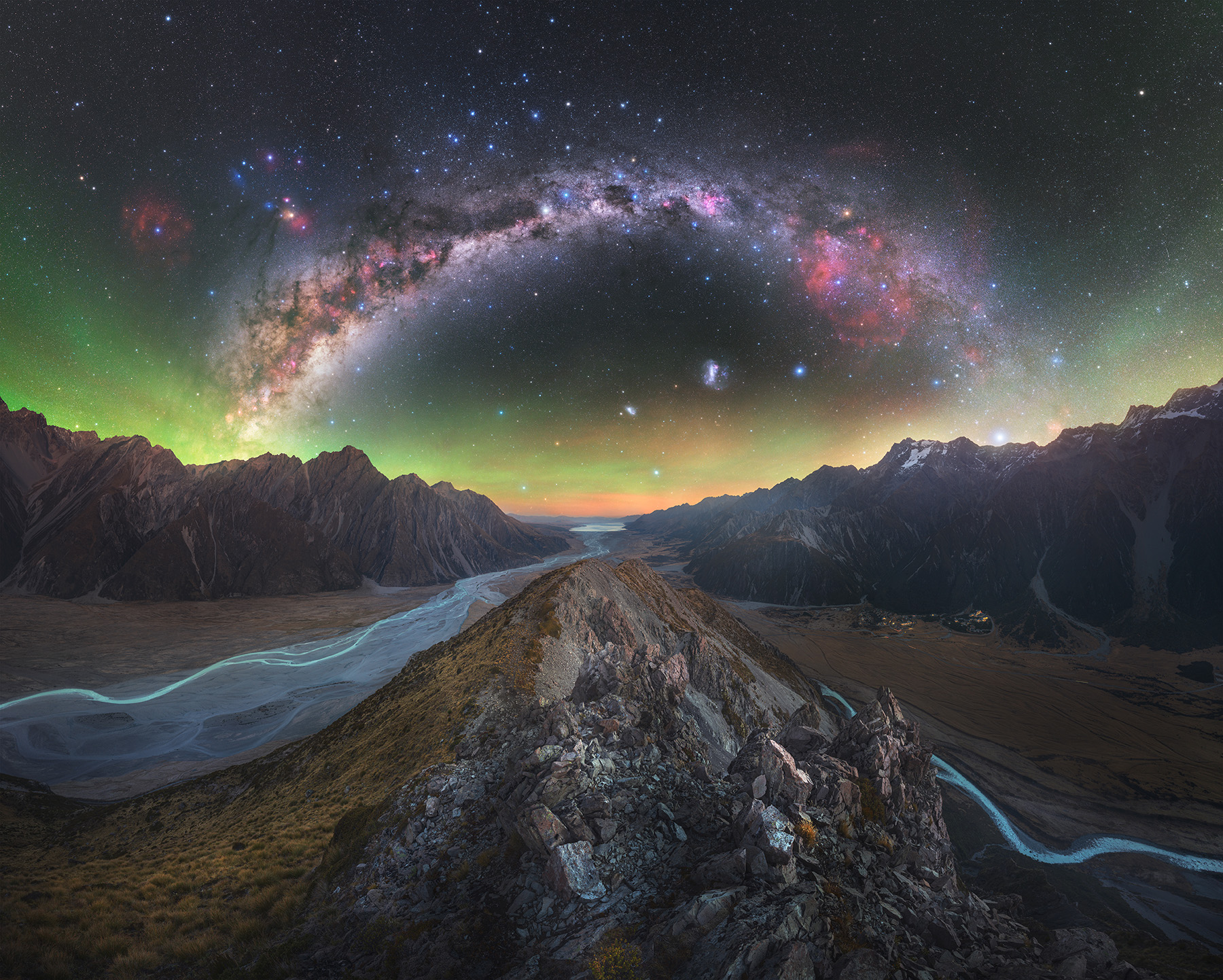 The glowing band of the Milky Way is pictured streaming in an arc across the night sky above a pair of meandering streams threading through a rocky landscape.