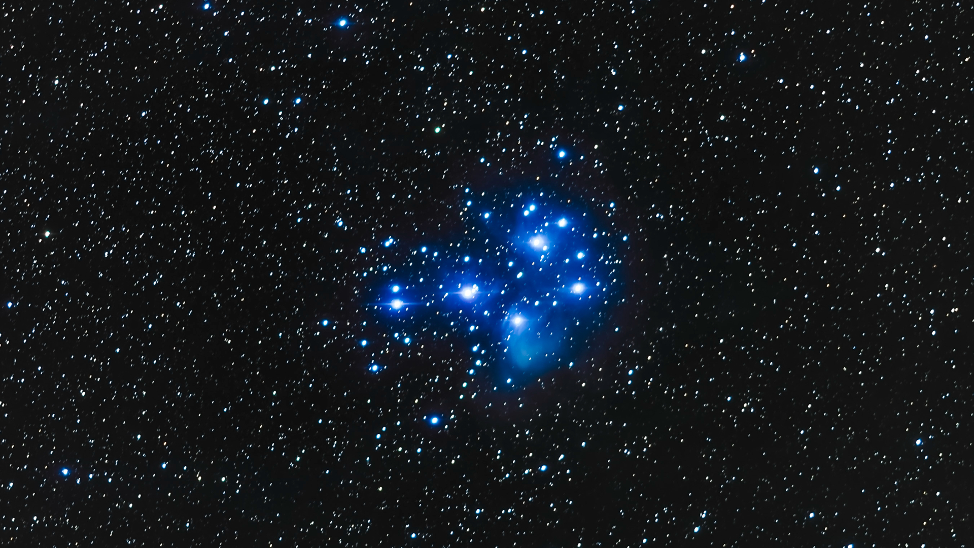 A collection of bright blue glowing stars is pictured in the center of an image hanging against a dense starfield of lower-magnitude stars framed by the blackness of space.
