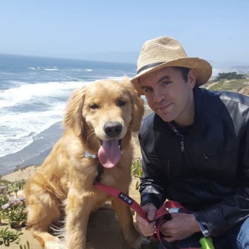 Man with wide-brimmed hat crouching down with his golden retriever dog, with the ocean in the background.