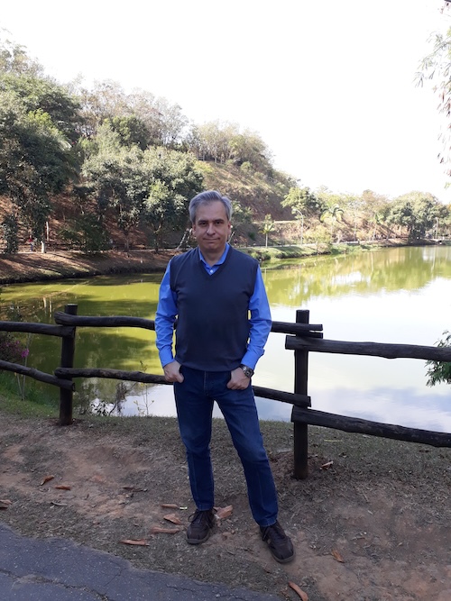 Man wearing dark blue jeans, dark vest and blue shirt stained beside a wooden fence, with a lake behind him.