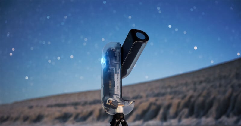 A modern, compact telescope with a transparent casing stands on a tripod, aimed at a star-filled night sky, with a rocky, blurred landscape in the background.
