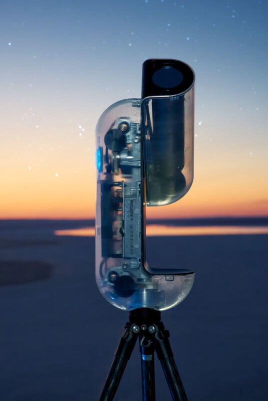 A modern, transparent telescope on a tripod stands outdoors at dusk, with a glowing sky and scattered stars visible in the background.