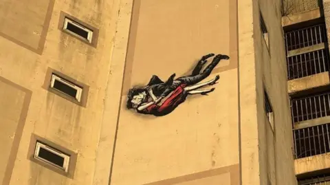 BBC An artwork of a man in a dark suit kissing a woman in a red dress and red heels as the y fall through the air. The art is on the side of a concrete building. It is bathed in golden sunlight.