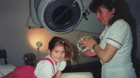 Family photograph A four-year-old Kat Watson-Wood in hospital wearing her hair in a high pony wearing a white shirt and hot pink skirt smiling. A nurse with long brow curly hair smiles looking at the youngster. 