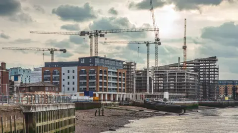 Getty Images Several cranes stand over riverside construction sites, where new apartment blocks and modern buildings are being built. In the foreground, a moored barge rests on the riverbank beside the water, while scaffolding and partially completed structures dominate the skyline under a cloudy sky.
