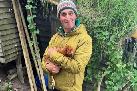 Dave Jacobs Dave Jacobs smiling at camera and holding a chicken. He is wearing a mustard-coloured jacket and a multi-coloured wooly hat. There is greenery behind him and a wooden structure.