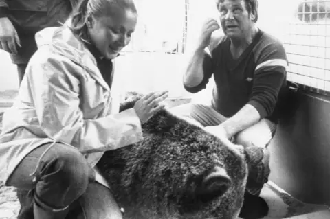 PA Media A black and white photo of a woman, man and a bear. The woman is kneeling and petting the bear, while the man sits nearby, facing the camera.