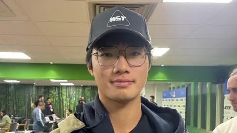 James Patient/BBC Xianghao Xiao wears a beige jacket over a black hoodie, and has a black cap on his head. He is standing in a media room at the Brentwood Centre. The room has green walls. Various people are sitting or standing in the background.