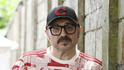 Matt Eccles Matt Eccles has a dark moustache and is wearing a black cap, black framed glasses and a red and white T-shirt. He is leaning against an outside wall.