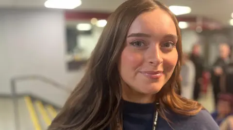 Rhianna Venables/BBC Elsie Booth has long brown hair and is smiling while wearing a navy top.