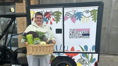 The Courtyard Pantry A female volunteer, with glasses and a light green top, holds a basket of vegetables while standing outdoors next to an electric quadricycle, saying THE COURTYARD PANTRY ENTERPRISE on it.