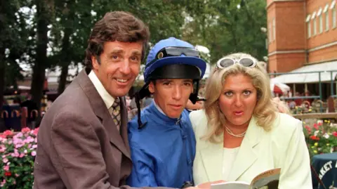 PA Media John Stapleton, wearing a brown suit, smiles alongside jockey Frankie Dettori, wearing blue silks, and Vanessa Feltz at Newmarket race course