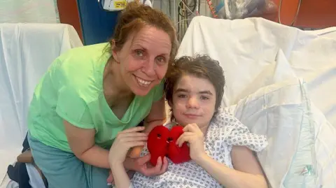 Family A woman wearing a green shirt and pale green trousers, smiles as she stands next to a boy with dark hair who is in a hospital bed wearing a gown. They are both holding cuddly toy kidneys. 
