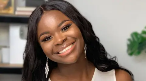Temi Danso A smiling woman with long black hair and a white top, standing in front of a white background with the blurred shape of shelves on the wall behind her