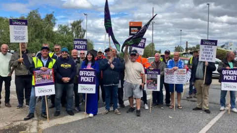 Nicola Rees/BBC About 20 staff members on the picket line holding up signs with Unison's purple branding. They are mostly in casual clothing, many seemingly in their 60s.