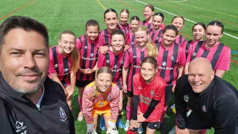 Colin Smith A team photo with two coaches and a dozen girls wearing black and pink jerseys