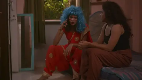 Flip Films Two women in a dimly lit apartment - one wearing a black vest, the other wearing an ornamental red saree with a blue wig, talking on the phone.