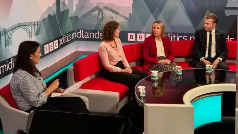 Presenter Elizabeth Glinka and three est Midlands MPs are sitting on a red sofa in the BBC Politics Midlands studio. There is a curved table in front of them, with four mugs on it. There is BBC Politics Midlands branding on a backdrop behind the sofa.