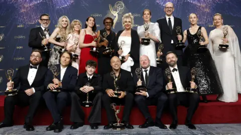 EPA Sixteen people, split into two rows, pose for a picture in front of a purple wall decorated with the Emmys logo. Six of them are seated at the edge of a stage covered in red carpet while the others stand behind them. They are all smartly dressed and hold golden trophies in their hands as they smile for the camera. 