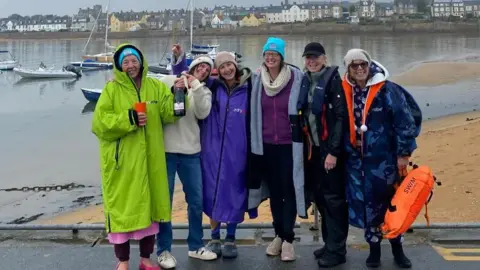 Six women in dry robes and warm hats smiling broadly at the camera. The woman on the far left has a bottle of fizz. They are posing at the water's edge. Small boats and harbourside houses can be seen in the background. 