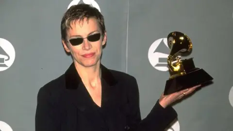 Getty Images Annie Lennox, holding up a Grammy award in her right hand, with short dark hair, wearing dark glasses and a black top and jacket. She is standing by a Grammy sign. 