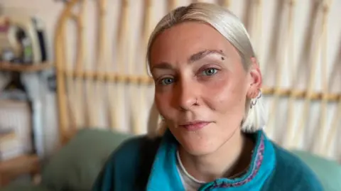 BBC Elle Glenny, who has bobbed blond hair, looking directly at the camera and wearing a turquoise fleece. There is a bed frame in the background.
