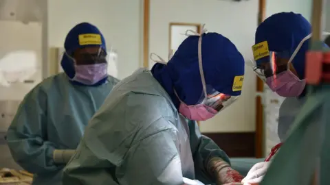 Somerset NHS Foundation Trust A group of people wearing medical scrubs and hats, with yellow name badges on. They're looking over a table as they are performing surgery.