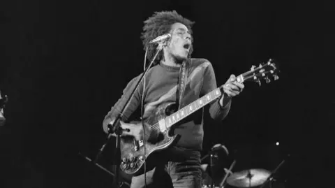 Gary Merrin A close up black and white image of Bob Marley in concert from 1973 clutching his guitar.