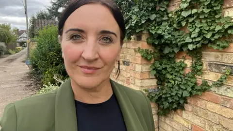 BBC Kath Sansom, founder of the patient-led campaign group Sling the Mesh, stood outside next to a brick wall. She is looking directly into the camera and has on a green blazer jacket over a black top. The wall behind her is covered in green ivy.