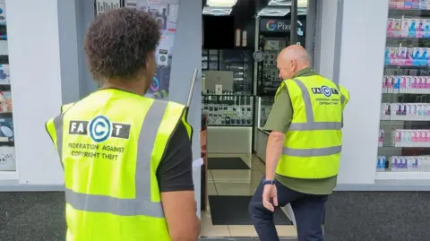FACT Two men wearing florescent vests with the Fact logo on the back are entering a shop front. There are boxes of vapes in one window and in the other there are electronics waiting to be resold.