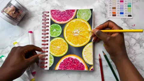Temi Danso A woman's hands holding a coloured pencil with a drawing of slices of citrus fruit on a notepad in front of her on a white marble surface, with other coloured pencils lying beside it