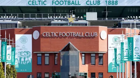 PA Media A view of the entrance to Celtic Football Club. It is a redbrick building with a walkway flanked by tall vertical flags showing players and slogans in green and white. A sign at the front top of the building says Celtic Football Club in silver and a gold star sits above the words.