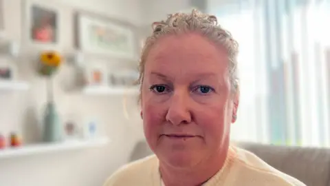 BBC Tracy Marelli is sitting on her cream sofa. She is wearing a yellow jumper, has blue eyes and blonde hair. Behind Tracy is a wall of tributes to her daughter from a blue vase filled with sunflowers to family pictures and various ornaments. 