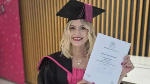 Sarah Gascoyne A woman wearing a black and pink university gown and cap at her graduation ceremony. She is holding her degree certificated, a white A4-sized piece of paper - in front of a black and gold background. 