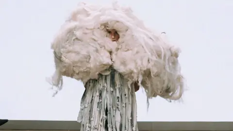 Gabrielle Thomas A model wearing one of Nathan Slate's designs on the catwalk. It is a dress made from rags dyed in different shades of grey and blue and tied into knots. Around their head is a large clump of white fluff and wool, designed to look like a mushroom cloud. 