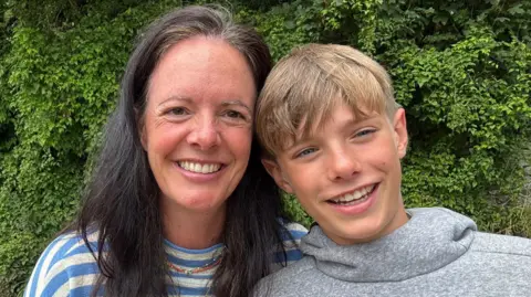 Sarah Sarah and her 11-year-old son Osian smiling at the camera. Sarah has long black hair and a striped white and blue top. Osian is wearing a grey jumper and has light brown hair.