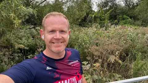 James McLaughlin A man in a blue and red sports t-shirt smiles at the camera as he takes a selfie. He has white airpods in his ears and stands in front of some bushes.