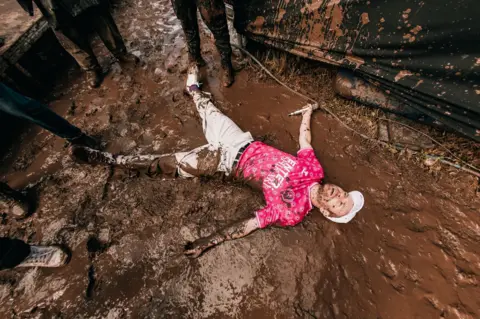 Jez Pennington Enter Shikari wearing a pink t-shirt and cream pants lies in a pool of mud while holding his mic in one hand