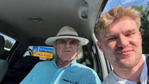 Driving Ukraine John Caldwell, sat in a vehicle, is wearing a white hat and Fynn Watt, is leaning through the vehicle's left front window, as they both pose for the picture with a sign for Ukraine visible through the vehicle's back windscreen.