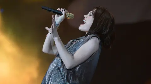 BBC Jessie J singing into a microphone, leaning back, on stage. She is wearing a glittery silver dress, has red lipstick, and has black hair.