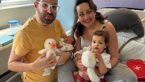Family picture A man and a smiling woman are seated on a hospital bed. The man is holding a newborn and a duck toy, the woman has on her lap a toddler.